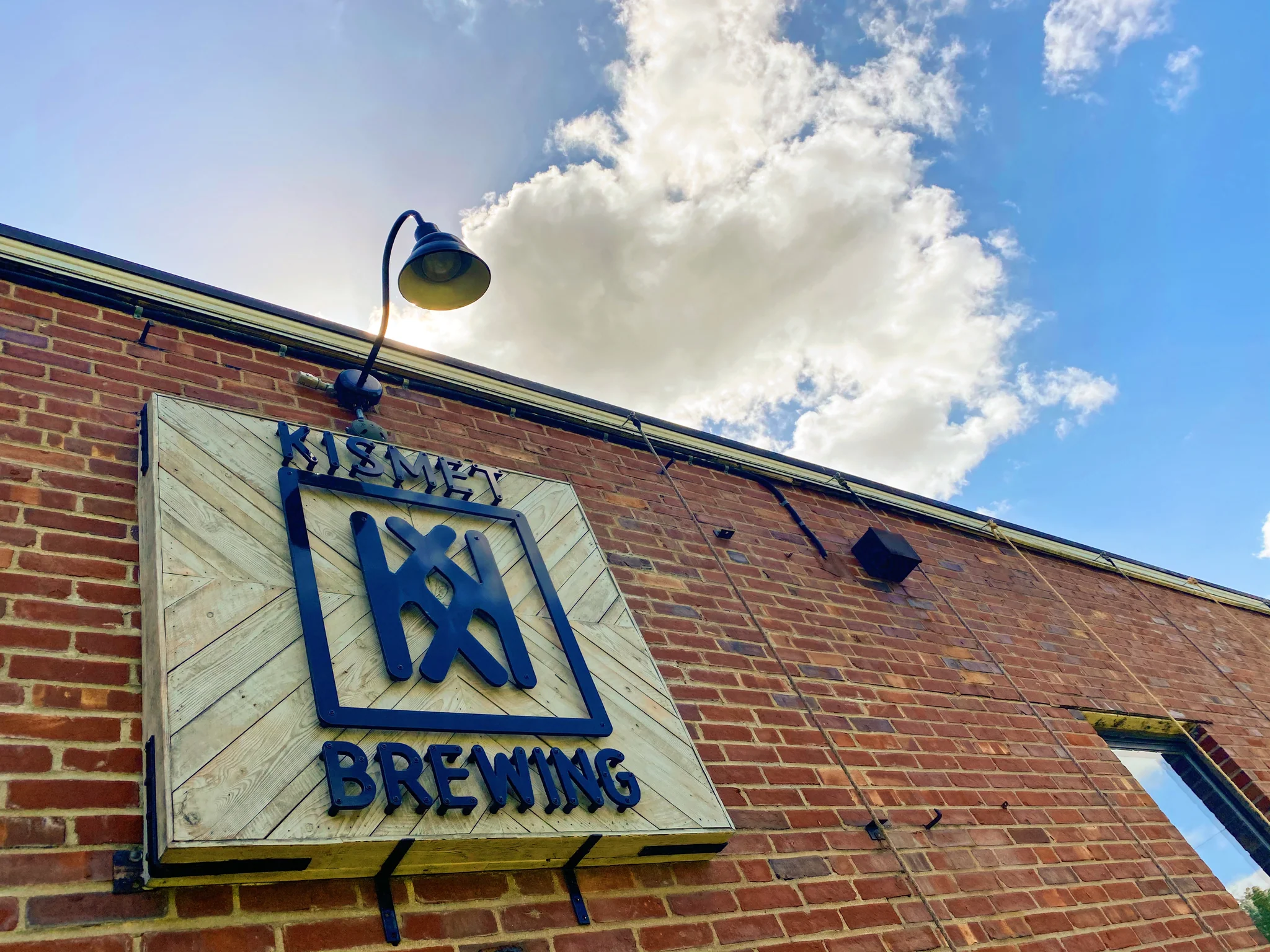 a low angle view of the sign for Kismet Brewing feauring the logo beneath a lamp, the sign hangs on the brick exterior of the brewery, a cloud hangs in the blue sky above