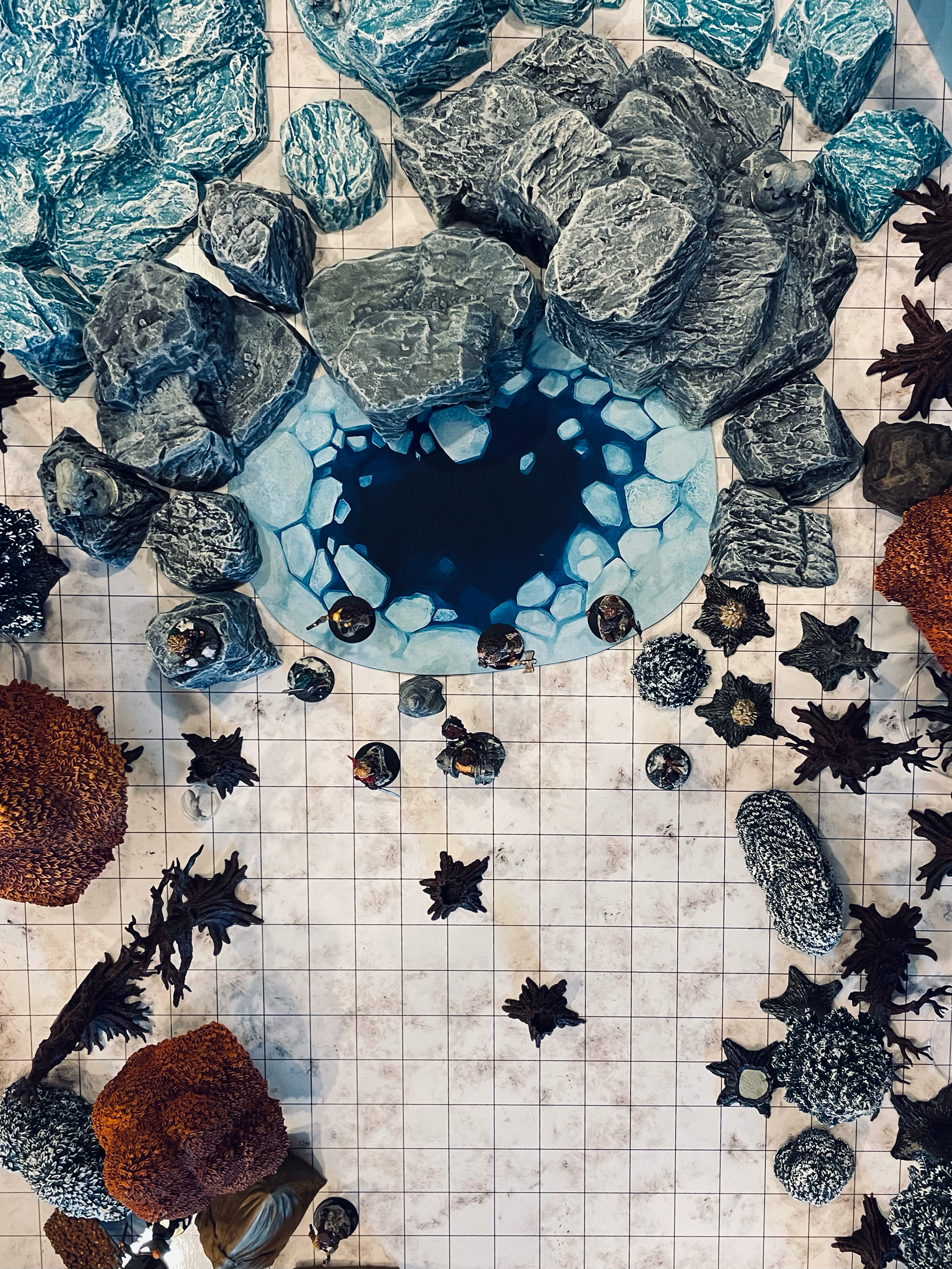 an overhead view of a winter wilderness diorama from a D&D game featuring figures at the edge an icy pond surrounded by rocky outrcroppings